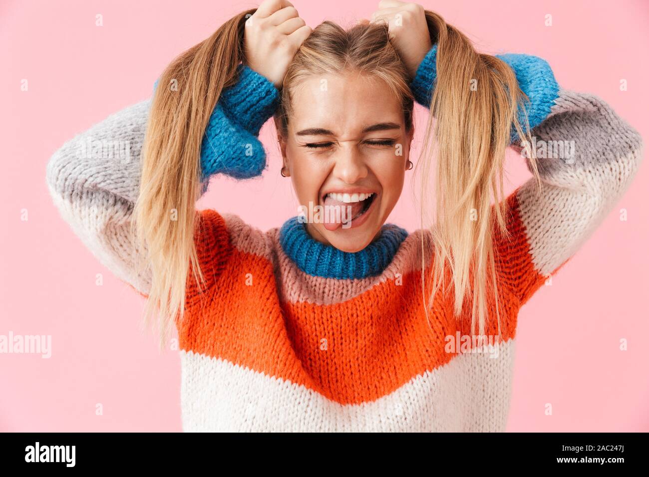 Portrait d'un drôle de jeune fille blonde wearing sweater s'amusant plus isolé sur fond rose, tirant sur ses cheveux Banque D'Images