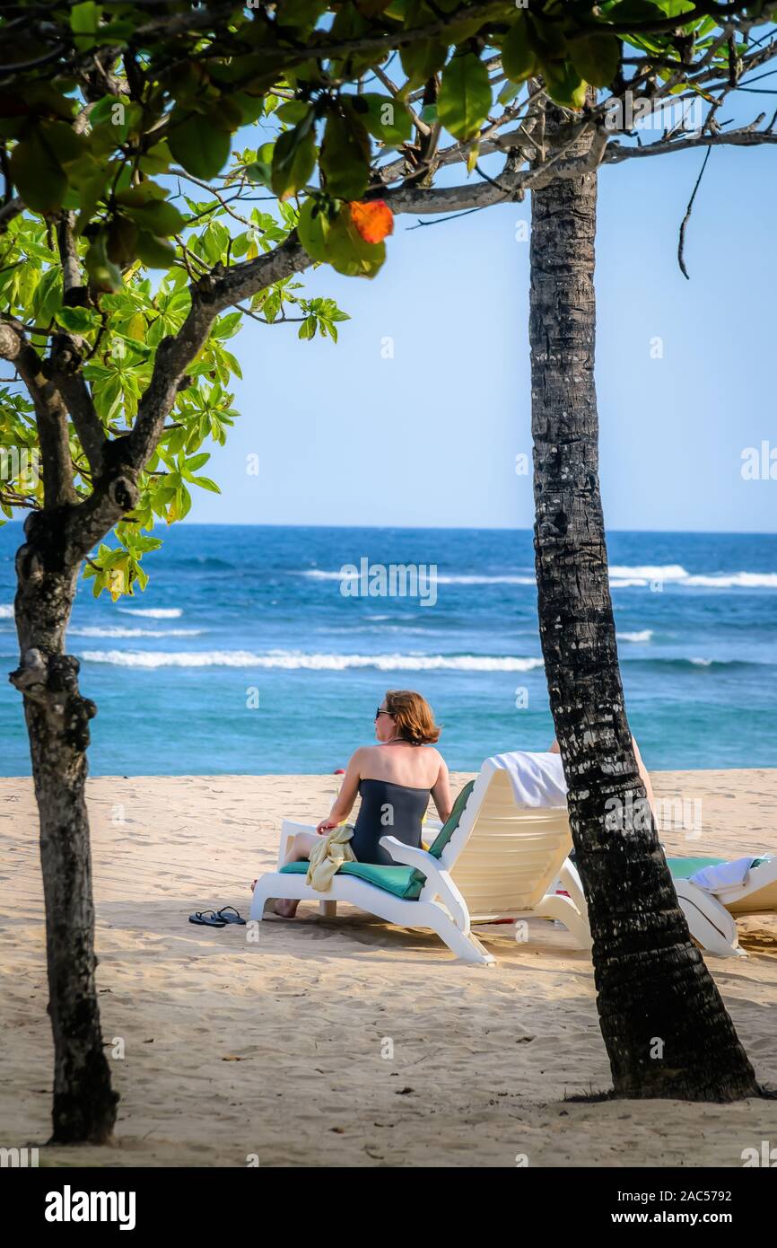 Une femme en maillot de bain assise sur une chaise de salon de plage bain de soleil sur une plage Banque D'Images