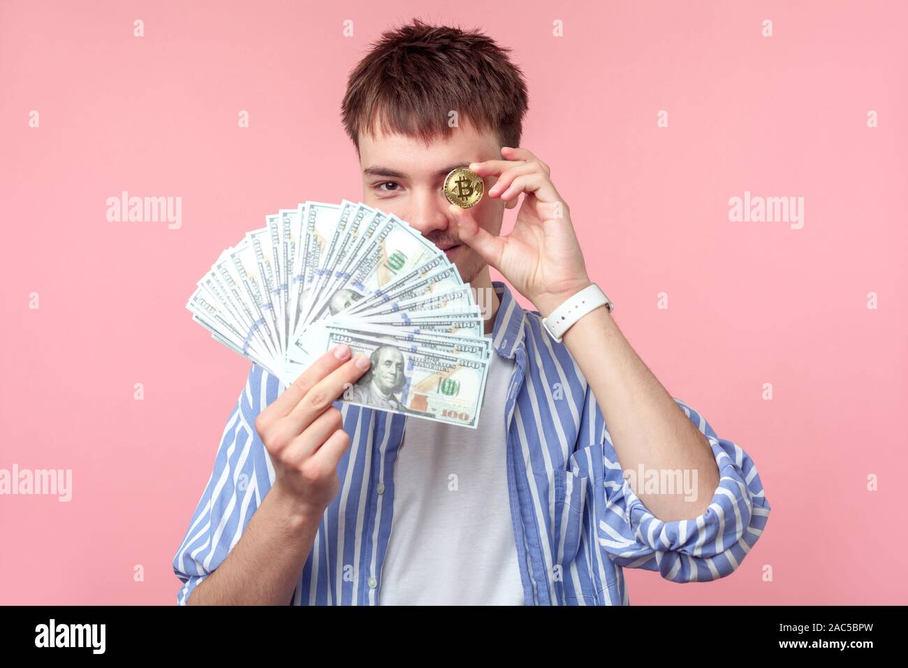 Portrait de jeunes homme aux cheveux brun avec petit barbe et moustache en chemise rayée décontractée avec un œil et bitcoin holding dollars. Banque D'Images