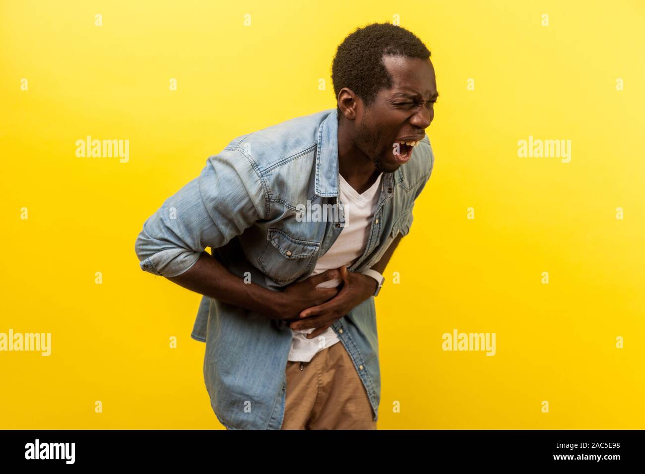 Portrait d'homme malade dans la chemise en jean penché et grimaçant d'une forte douleur abdominale, tenant les mains sur le ventre, ayant l'appendicite gastrite ou méthode sympto Banque D'Images