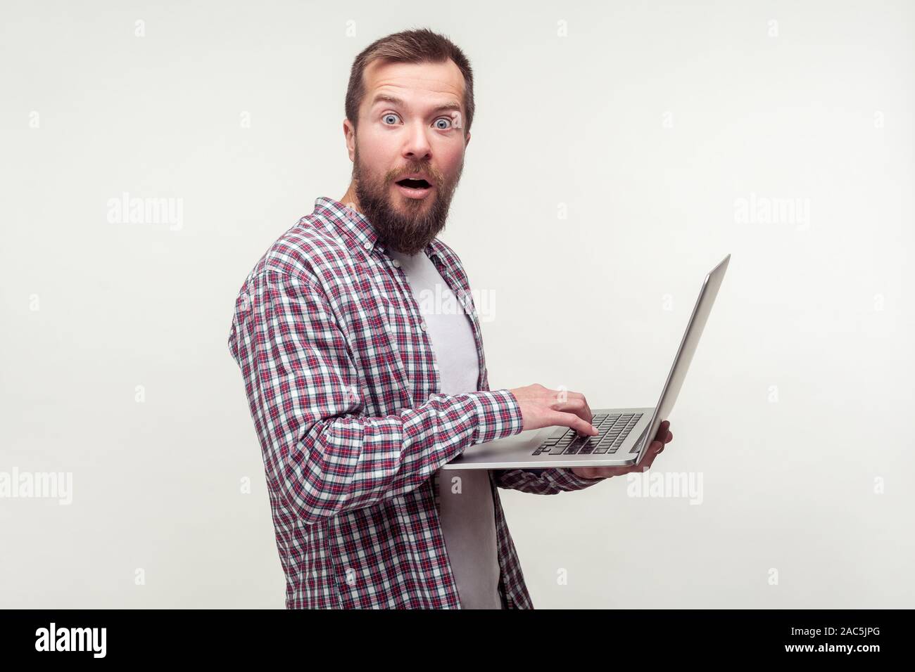 Portrait d'utilisateur d'ordinateur étonné, barbu en chemise à carreaux décontractée avec ordinateur portable et accès permanent à la recherche à l'appareil photo avec expression choquée, excité surpri Banque D'Images