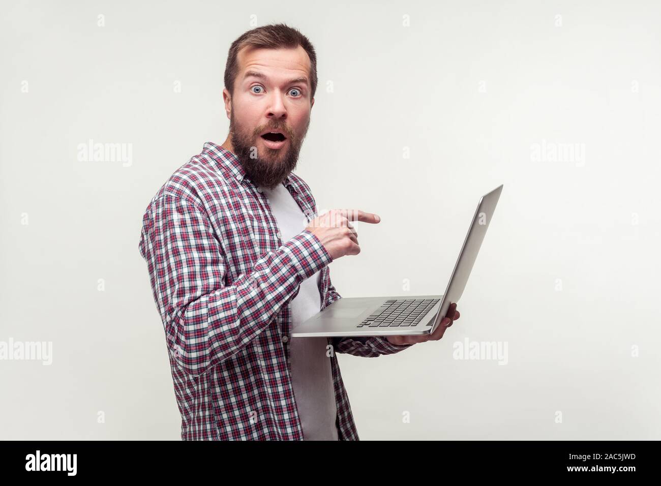 Portrait d'utilisateur d'ordinateur choqué, barbu en chemise à carreaux décontractée pointing at laptop, à la surprise d'application informatique, incroyable dans Banque D'Images