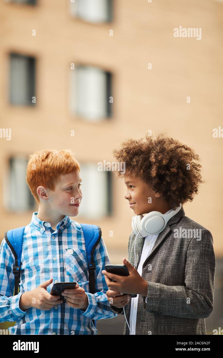Deux garçons souriants et permanent de parler les uns aux autres et à l'aide de leurs téléphones intelligents en plein air Banque D'Images