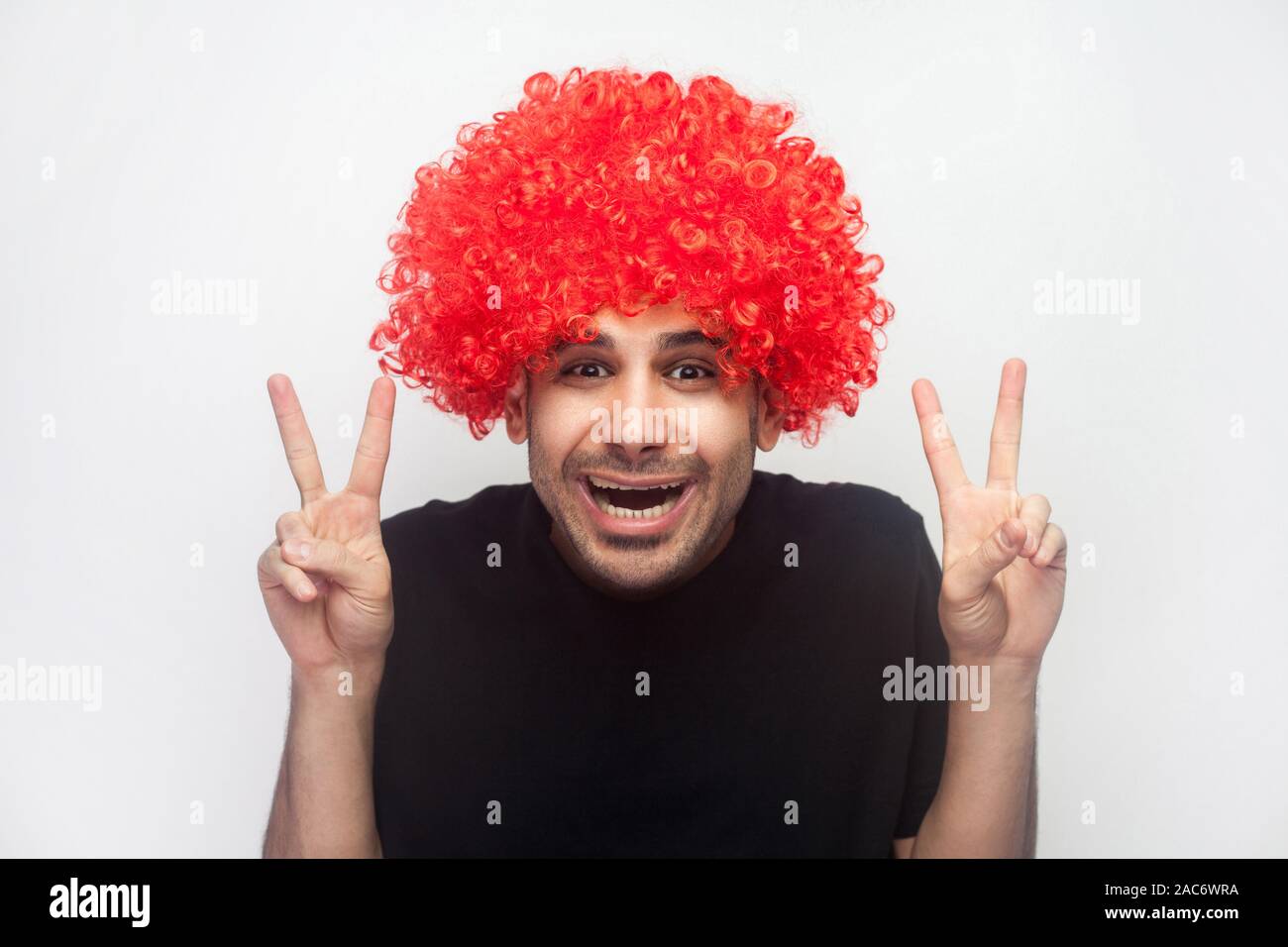 Portrait d'homme en soie avec hipster amical et bouclés perruque rouge montrant le double v signer la paix, la victoire ou geste de la main et hésitant. timide drôles Banque D'Images