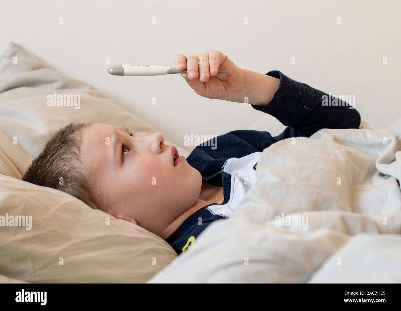 L'école maternelle garçon malade en pyjama couché sur un oreiller dans le  lit et en regardant le thermomètre numérique. Mauvais Garçon est la mesure  de la température du corps et ne se