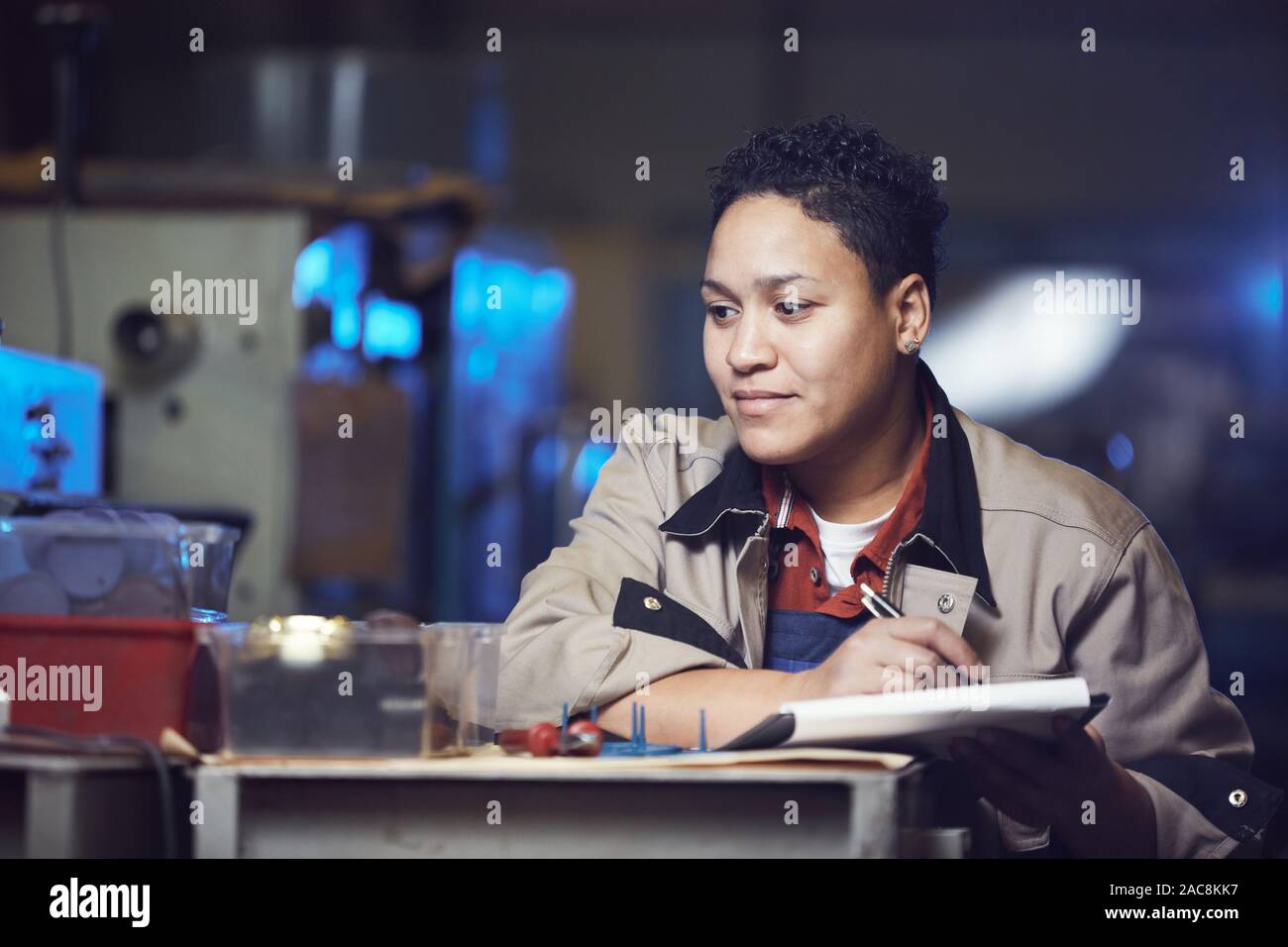 Portrait of smiling femme afro-américaine travaillant à la ligne de production à l'usine et prendre des notes sur le presse-papiers, copy space Banque D'Images