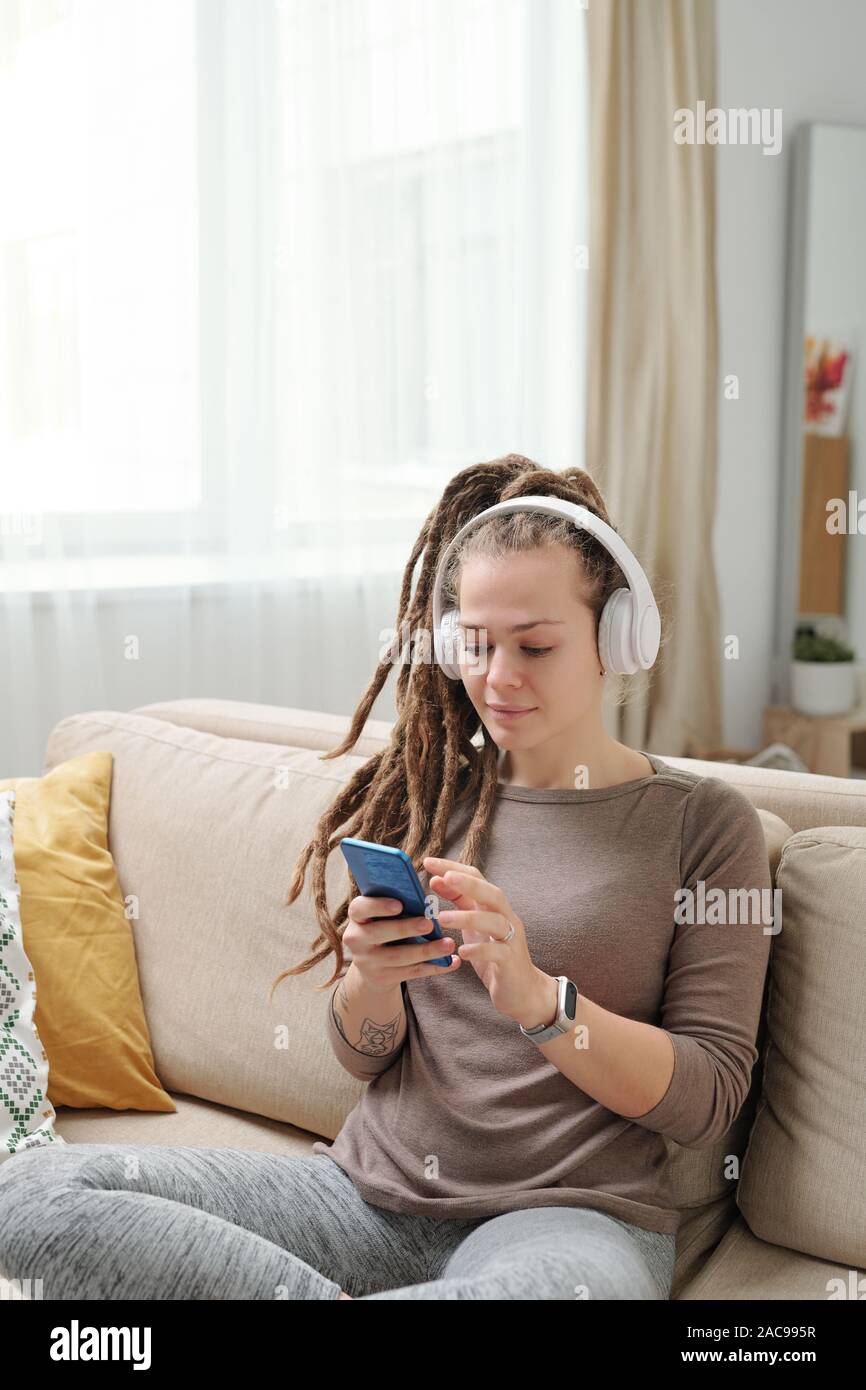 Jeune femme avec des dreadlocks défilement dans smartphone pendant que sitting on couch Banque D'Images