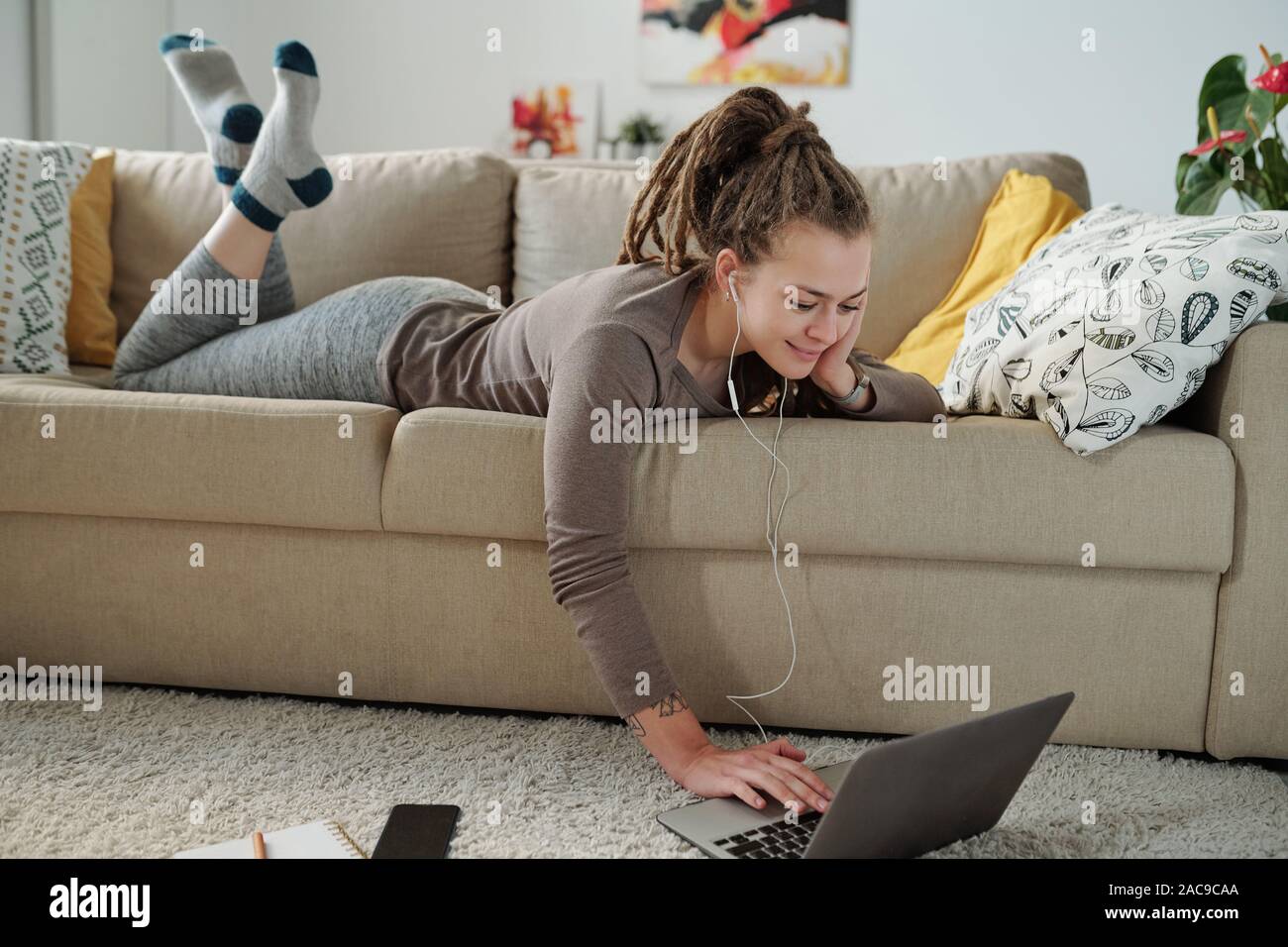 Smiling girl in earphones lying on sofa confortable et regarder des vidéos. Banque D'Images