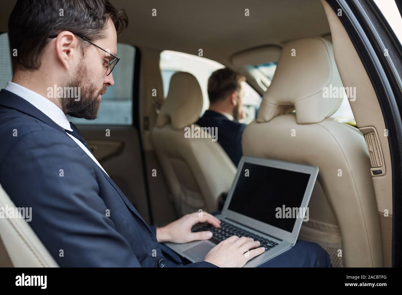 Young Woman in suit assis sur le siège arrière de la voiture et de travailler en ligne à l'aide de son ordinateur portable tout en conduisant la voiture pilote Banque D'Images