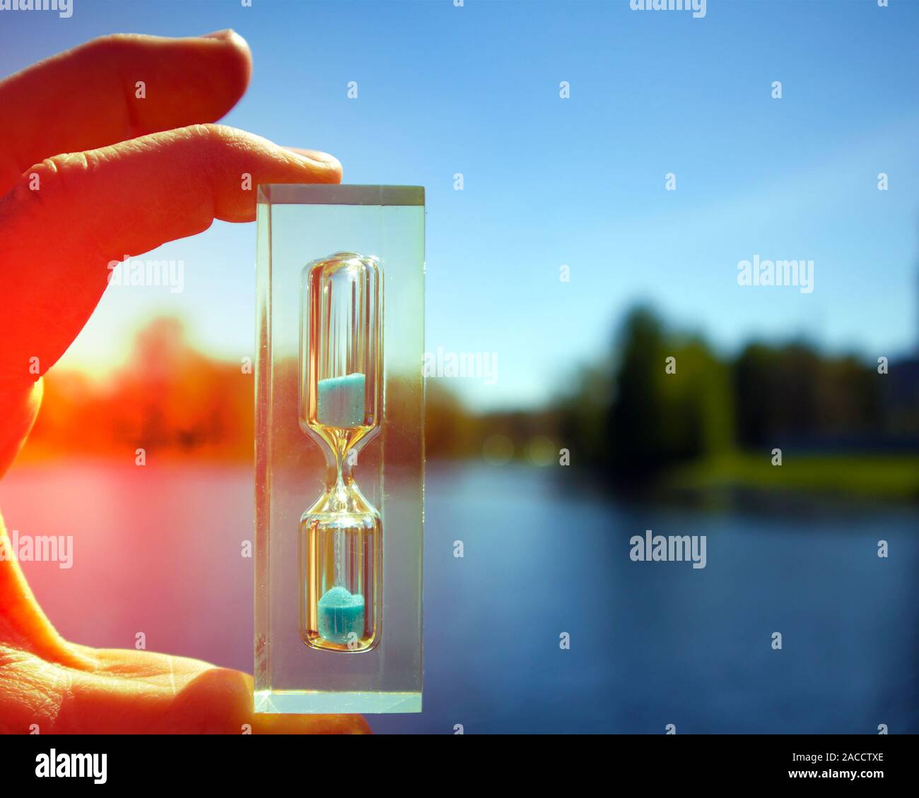 Sunlit human hand holding transparent avec hourglass sable bleu contre lac calme, concept de temps Banque D'Images