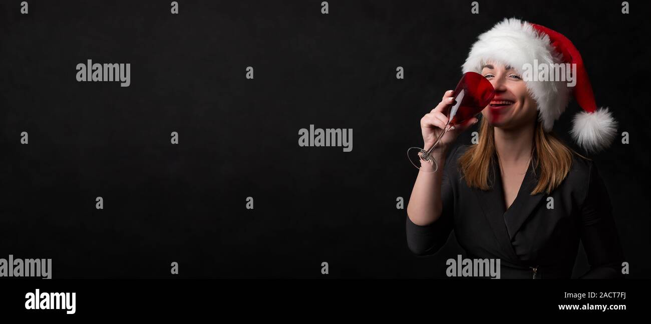 Une belle fille de santa hat boissons champagne à partir d'un verre de rouge sur un fond noir. Concept pour bannière, calendrier ou d'une carte de vœux dans happ Banque D'Images