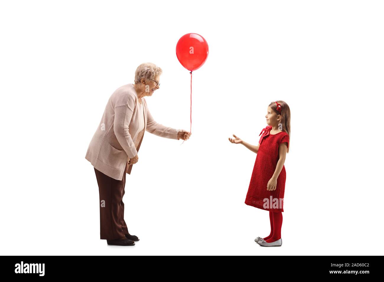 Profil de pleine longueur tourné d'une grand-mère en donnant un ballon rouge à une fille dans une robe rouge isolé sur fond blanc Banque D'Images