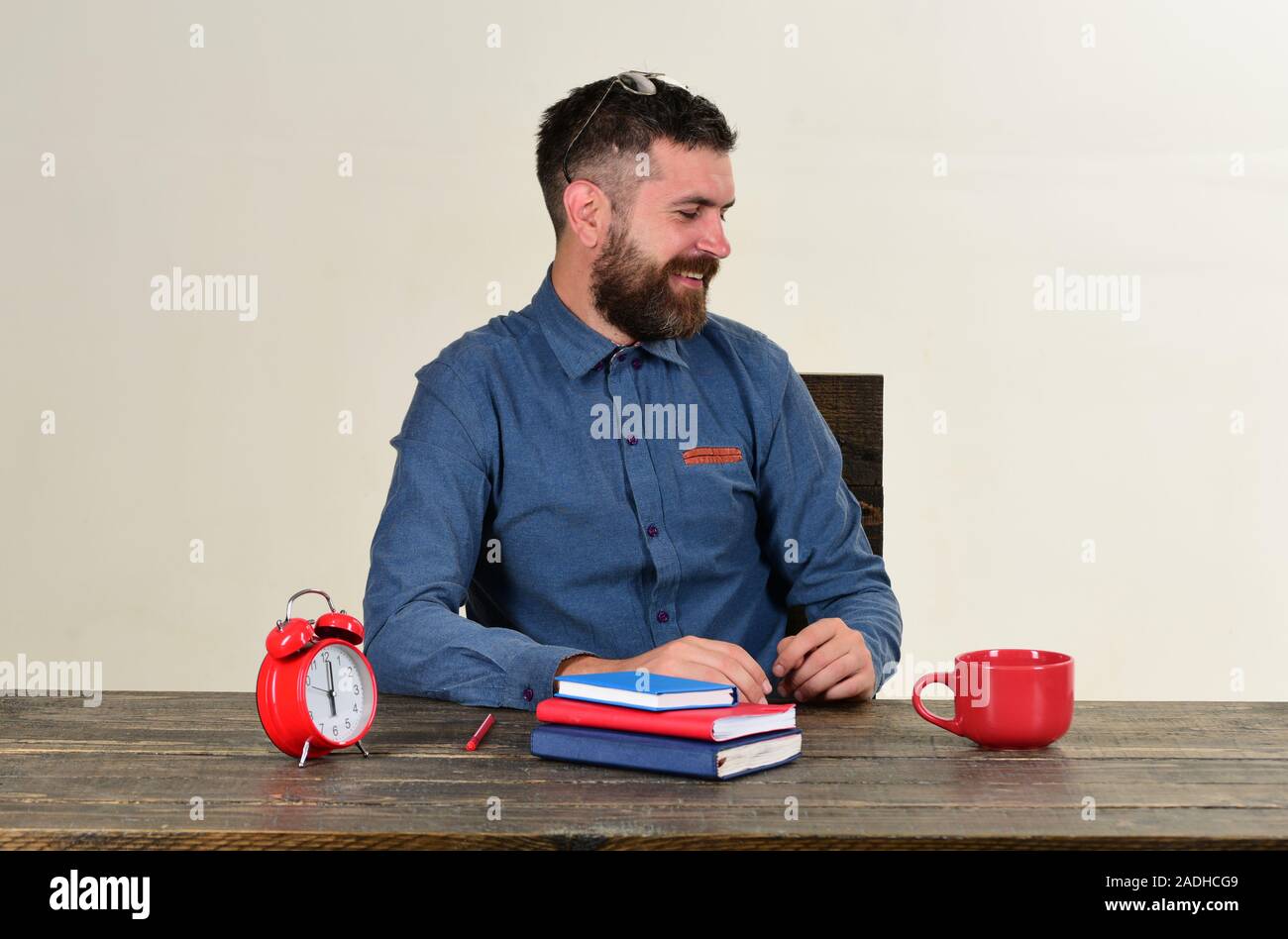 Homme à barbe est titulaire d'ordinateur isolé sur fond blanc. Examen et étude de concept. Avec des enseignants professionnels visage souriant est assis à un bureau en bois. Tasse et rétro avec horloge rouge et bleu livres sur table vintage Banque D'Images
