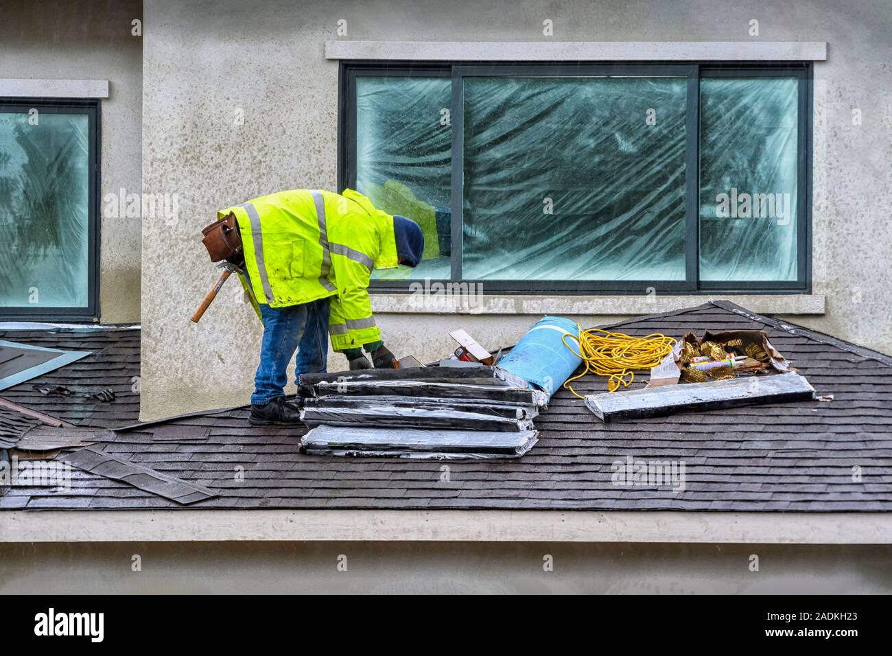 Notion de processus de construction. A Construction worer qualifiés l'ajout d'un toit en bardeaux d'asphalte sur une nouvelle maison, travailler sous la pluie. Banque D'Images