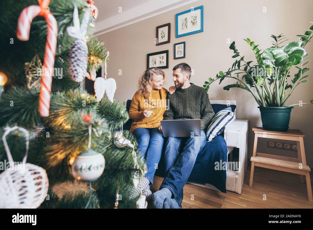 Young Caucasian couple shoppings de Noël en ligne sur le canapé à la maison avec un ordinateur portable, looking at each other and smiling Banque D'Images