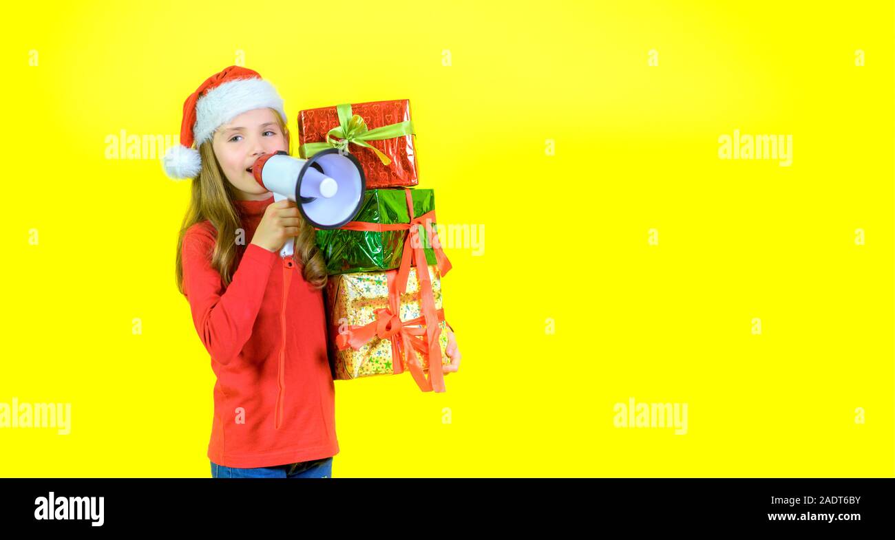 Girl in santa claus hat avec boîte boîtes dans la main crie dans un mégaphone, sur un fond jaune studio. La joie. Temps d'émotions Banque D'Images