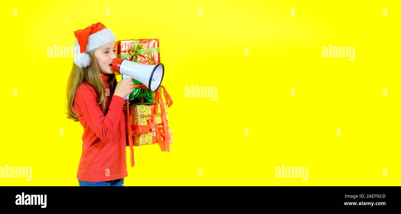 Girl in santa claus hat avec boîte boîtes dans la main crie dans un mégaphone, sur un fond jaune studio. La joie. Temps d'émotions Banque D'Images