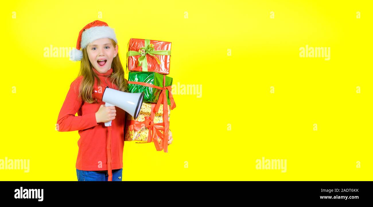 Girl in santa claus hat avec boîte boîtes dans la main crie dans un mégaphone, sur un fond jaune studio. Surprise. Temps d'émotions Banque D'Images