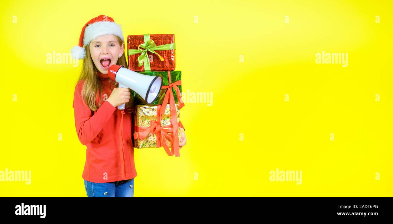 Girl in santa claus hat avec boîte boîtes dans la main crie dans un mégaphone, sur un fond jaune studio. Temps de vente Banque D'Images