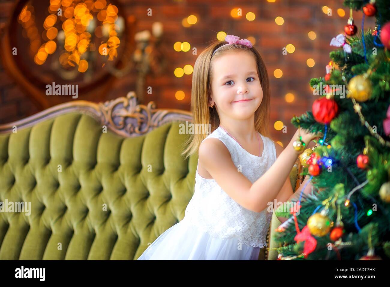 Jolie fille en robe blanche assise sur un canapé décore un arbre de Noël,  dans le contexte des lumières de fête lumineuse. Noël et Jour de l'Ye Photo  Stock - Alamy