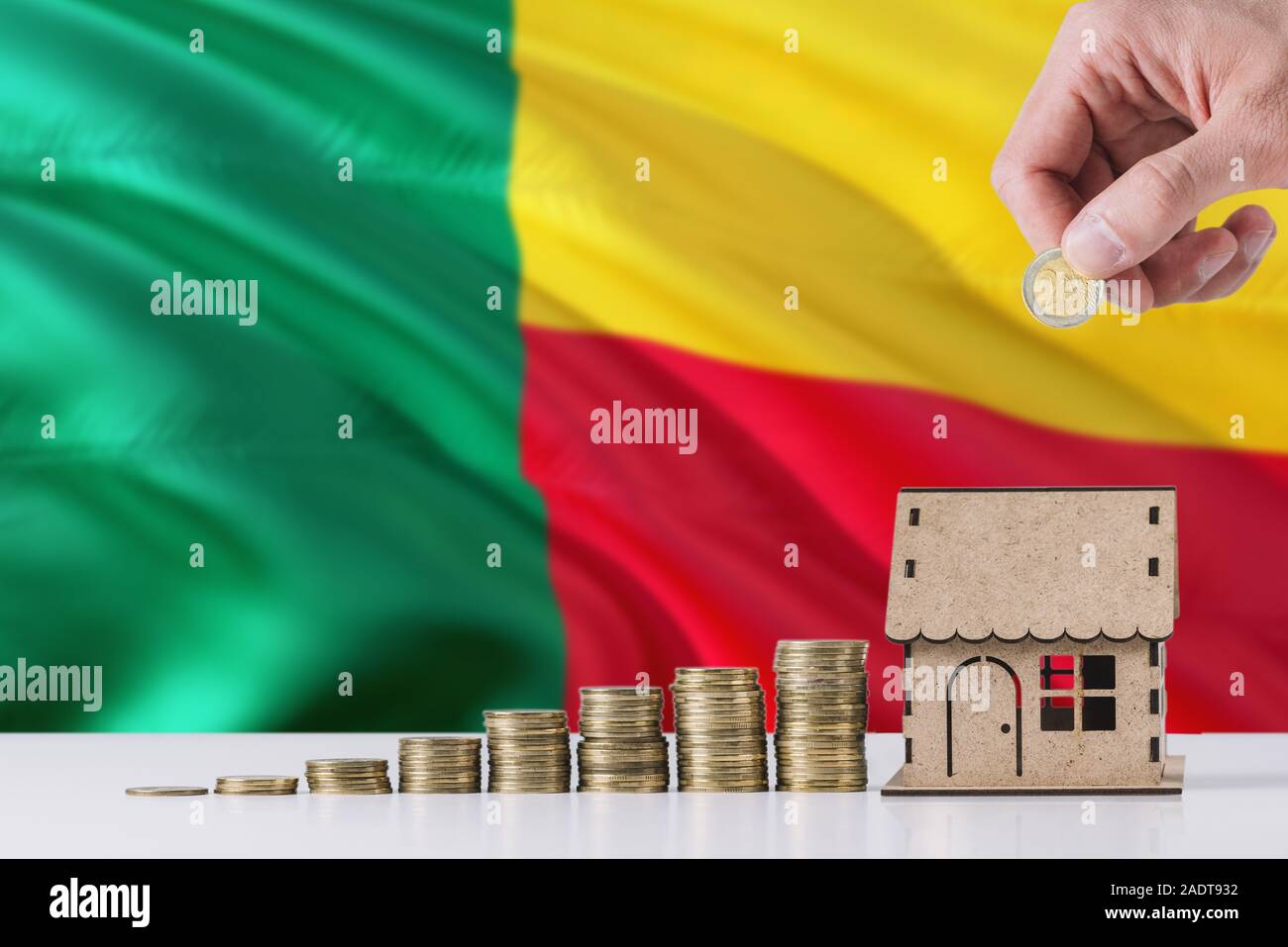 Man holding coins mise en maison en bois tirelire, de brandir le drapeau du Bénin dans l'arrière-plan. Économiser de l'argent pour prêt hypothécaire. Banque D'Images