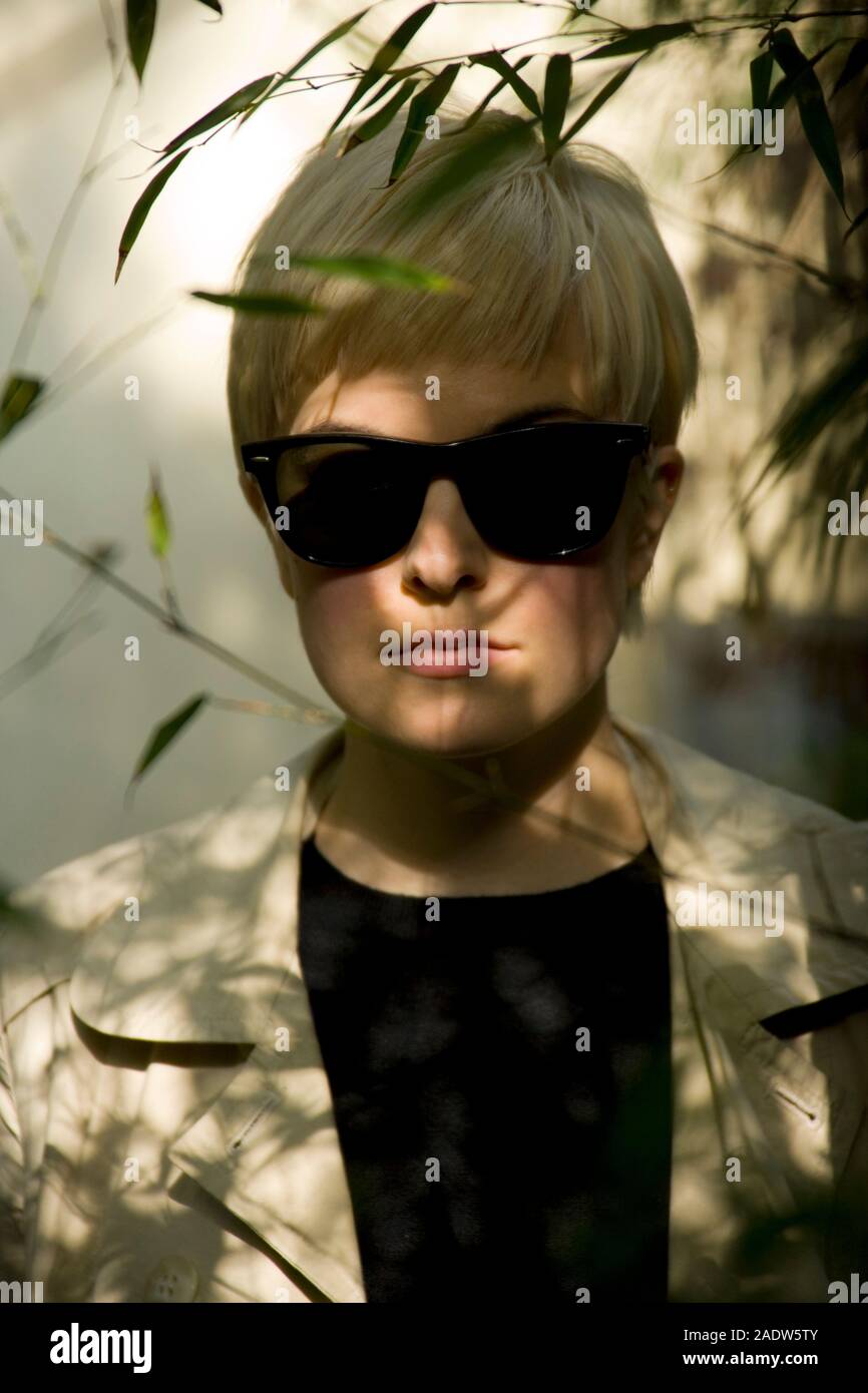Jeune femme élégante, 20's style des années 1950, le port de lunettes de soleil, imperméable et vu par le feuillage Banque D'Images