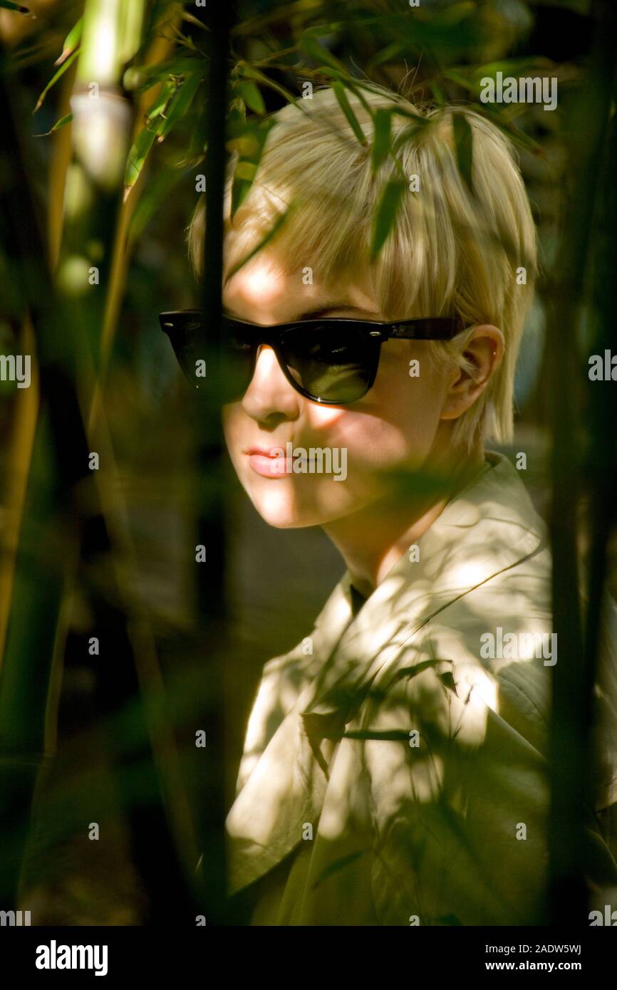 Jeune femme élégante, 20's style des années 1950, le port de lunettes de soleil, imperméable et vu par le feuillage Banque D'Images