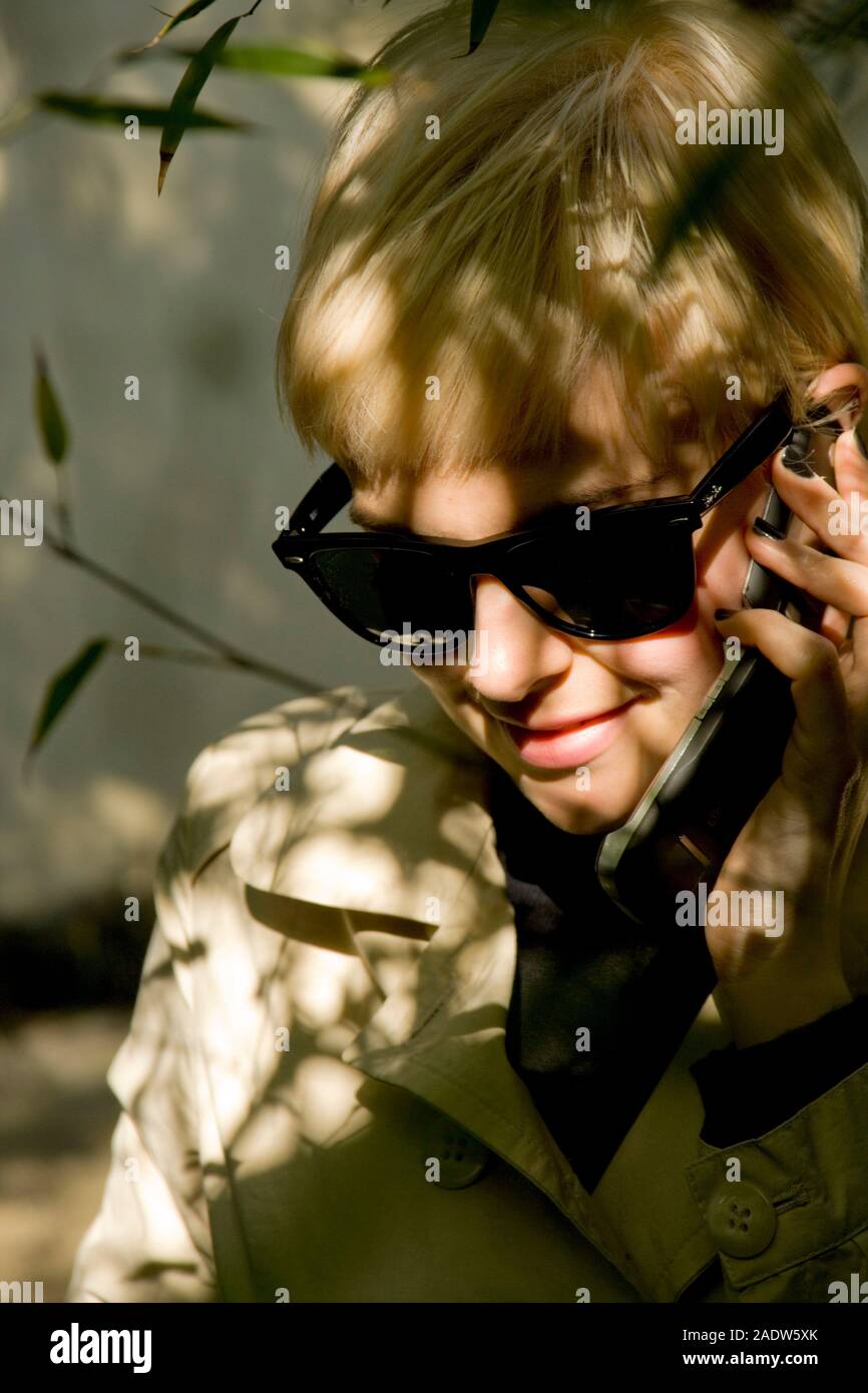 Jeune femme élégante, 20's style des années 1950, le port de lunettes de soleil, imperméable et vu par le feuillage à l'aide de téléphone mobile Banque D'Images