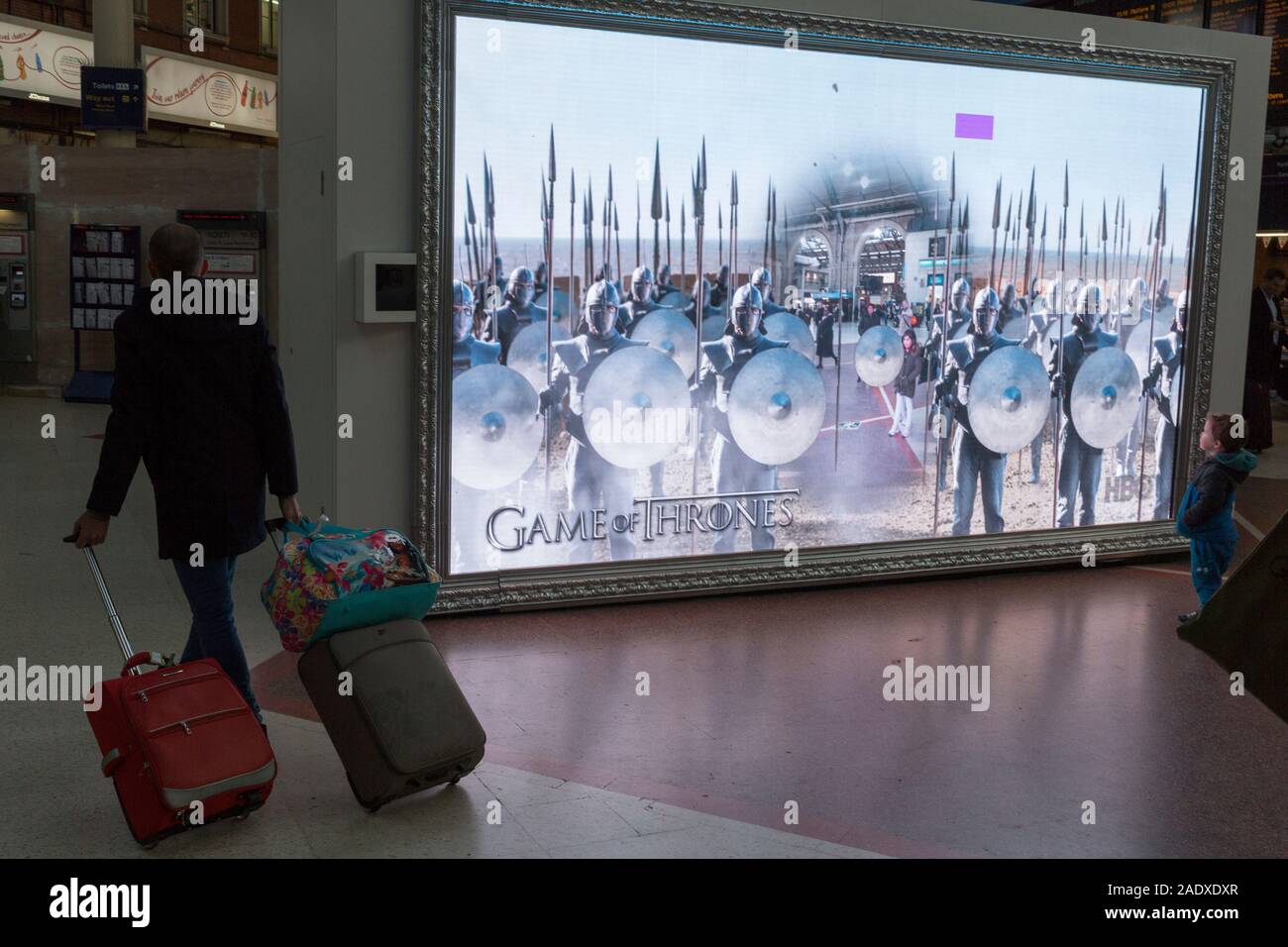 En tant que passager avec bagages marche dernières sur son chemin à une plate-forme, un jeune enfant regarde un grand écran montrant un monde de réalité augmentée du hall de gare et la fantaisie des créatures, partie d'une boucle de l'ads pour visiter l'Écosse et pour HBO's jeu des trônes, à Londres, en Angleterre, le 4 décembre 2019. Banque D'Images