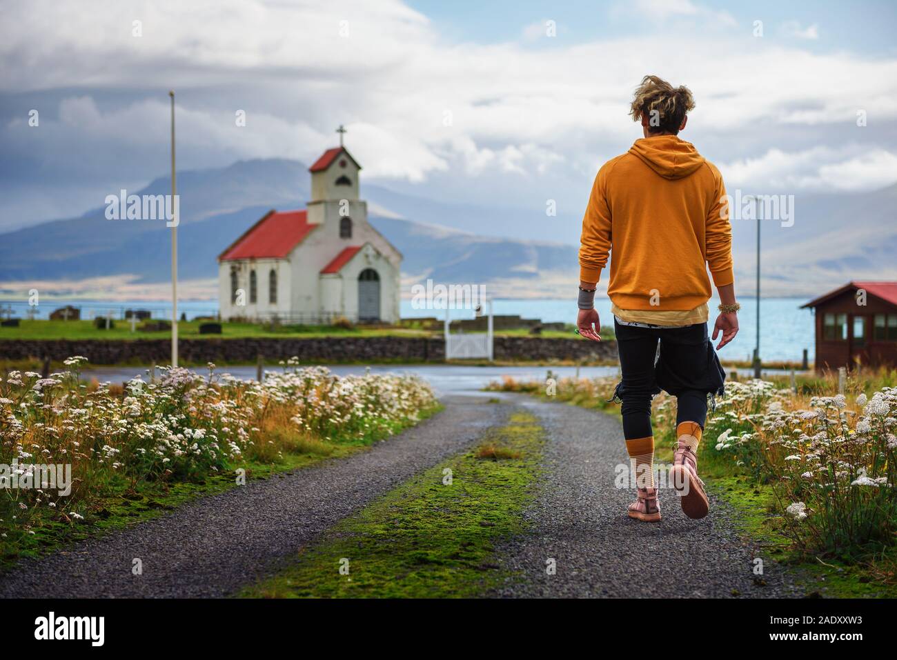 Jeune homme marchant sur un chemin de terre vers une église et un cimetière en Islande avec un lac à l'arrière-plan. La solitude, l'espoir, la tristesse , la dépression et Banque D'Images