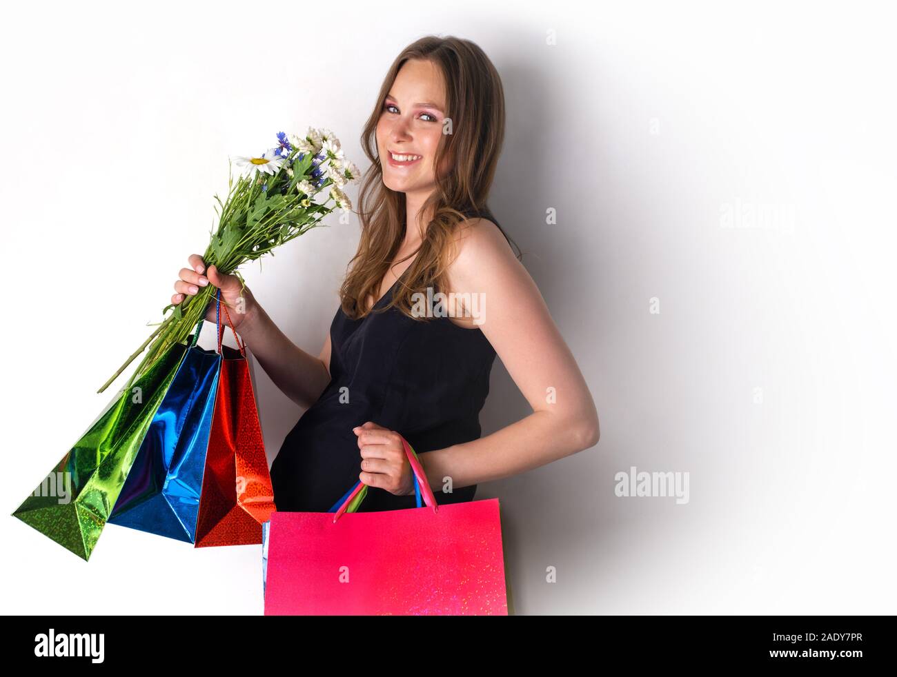 Une fille enceinte installée contre le mur avec des fleurs et les sacs du magasin. Joyful woman holding shopping bags bouquet de fleurs Banque D'Images