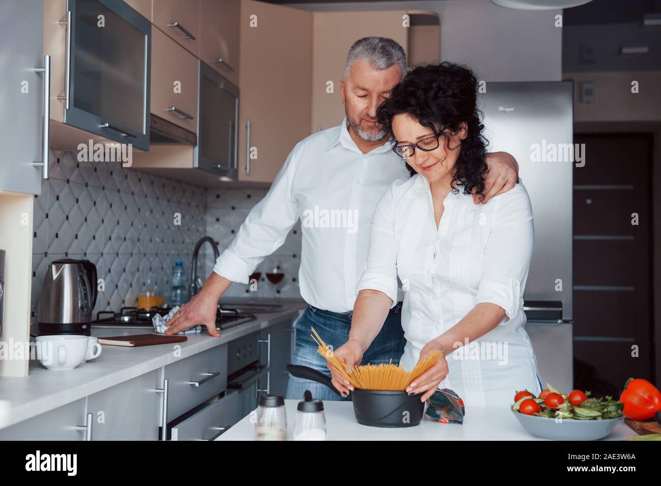 C'est ma recette secrète. L'homme et sa femme en chemise blanche à la préparation de la nourriture sur la cuisine en utilisant des légumes Banque D'Images