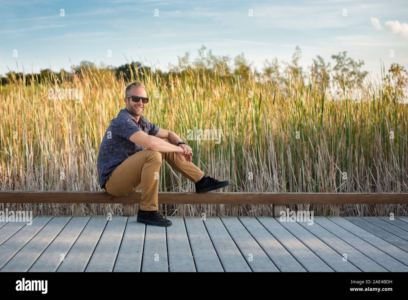 A smiling man in sunglasses est assise sur un trottoir en face d'herbes hautes Banque D'Images