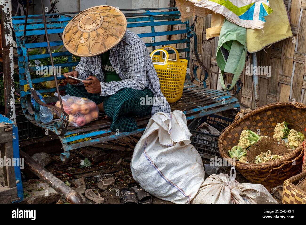 Un homme dans un chapeau de riz conique ou coolie hat se détend sur un banc bleu avec un téléphone cellulaire après l'achat de pommes dans le marché ferroviaire de Mandalay, Myanmar Banque D'Images