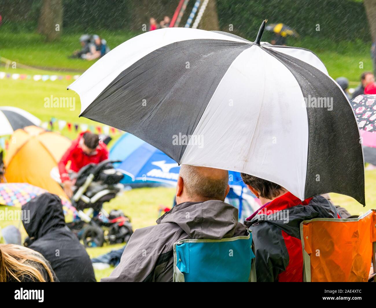 Couple sitting in replier sous de grandes chaises parasol à rayures regarder événement en plein air sous la pluie, Ecosse, Royaume-Uni Banque D'Images