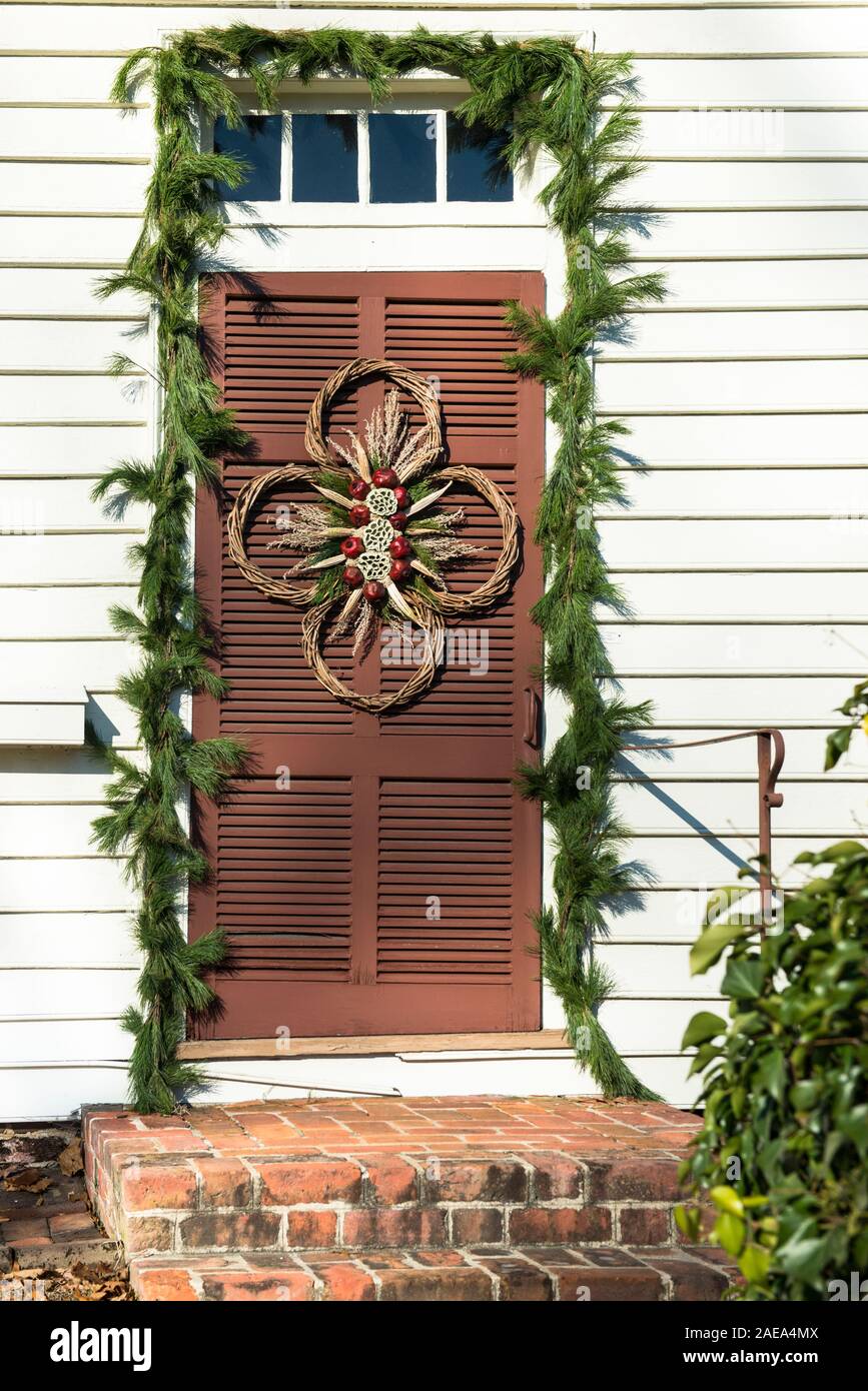Couronne de Noël décoration de porte, Colonial Williamsburg. Les gousses séchées Lotus rouge, grenades, bâtonnets torsadés, sur bois à la porte marron, evergreen swag. Banque D'Images