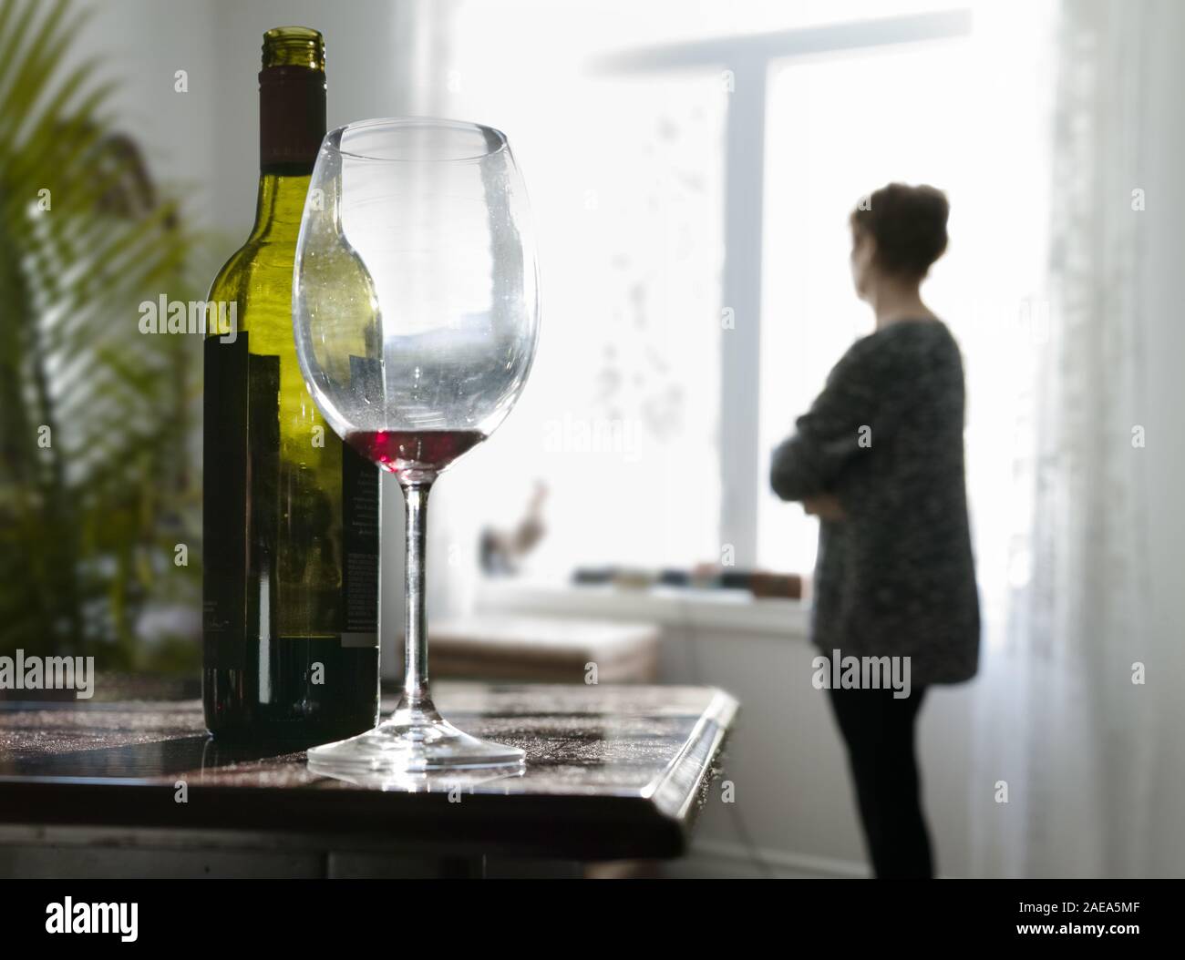 Une femme à l'arrière-plan, en regardant par la fenêtre, il est seul. Au premier plan, une bouteille de vin et un verre vide. Banque D'Images