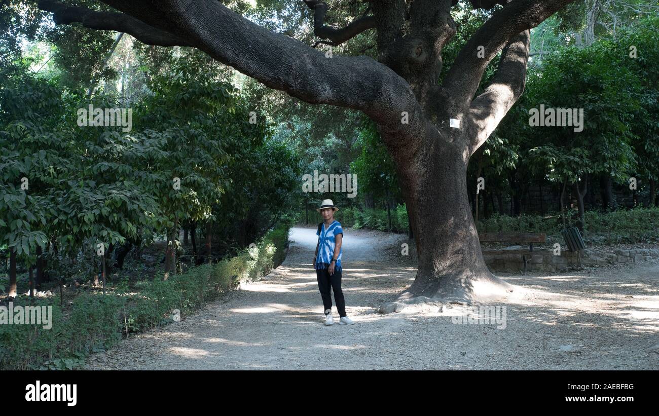 Asian woman with white fedora et cravate bleue-t-shirt mort debout par arbre dans les jardins nationaux à Athènes, Grèce Banque D'Images