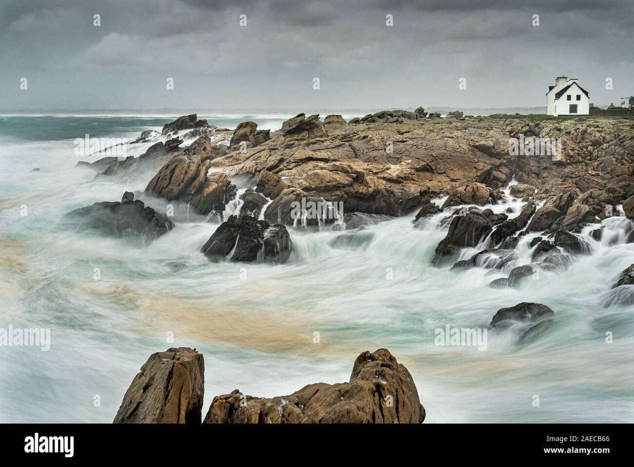 Une longue exposition à une tempête sur la côte de Bretagne. La mousse des vagues se brisant sur les rochers crée un brouillard blanc. Les maisons du village de stand Banque D'Images