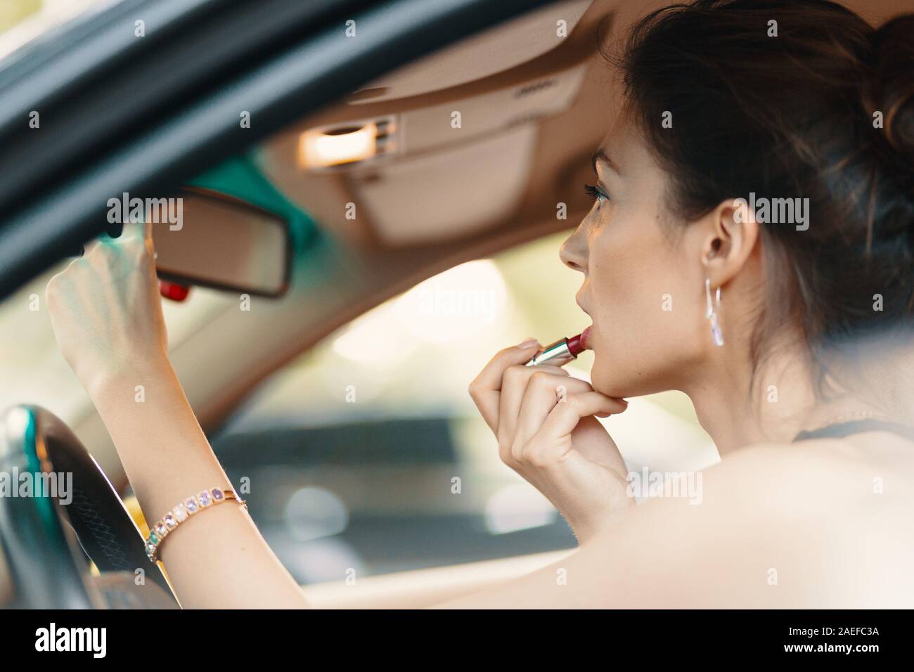 Jeune femme élégante à rétroviseur en peinture ses lèvres faisant l'application constituent en conduisant la voiture. Banque D'Images