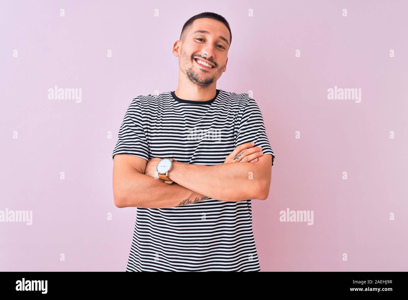 Beau jeune homme portant un t-shirt à rayures sur fond rose isolé permanent heureux face smiling avec bras croisés regardant la caméra. Positif Banque D'Images