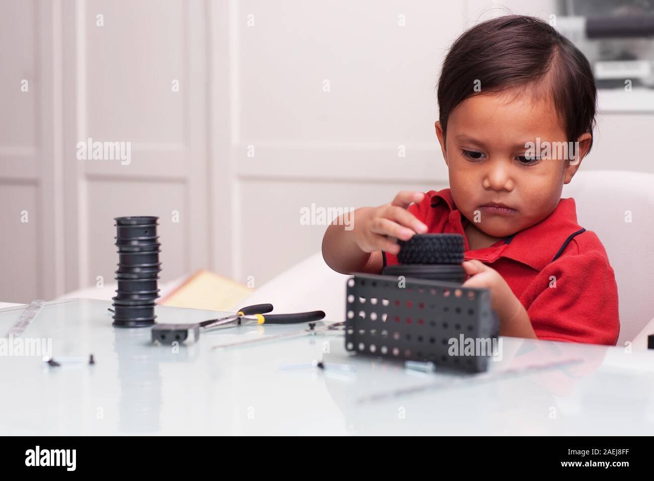 Un jeune garçon d'âge préscolaire, joue avec de petites pièces pour construire des tours à l'aide de la motricité fine sur une table en verre. Banque D'Images