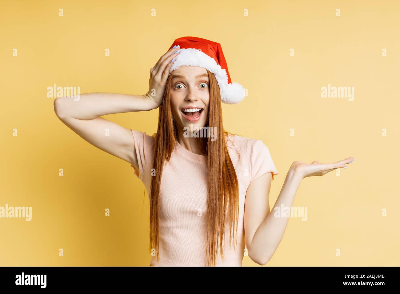 Stupéfait caucasian ginger girl in Santa hat et crème couleur t shirt, holding copy space, posant avec open palm, de montrer ou de présenter votre produit, ite Banque D'Images