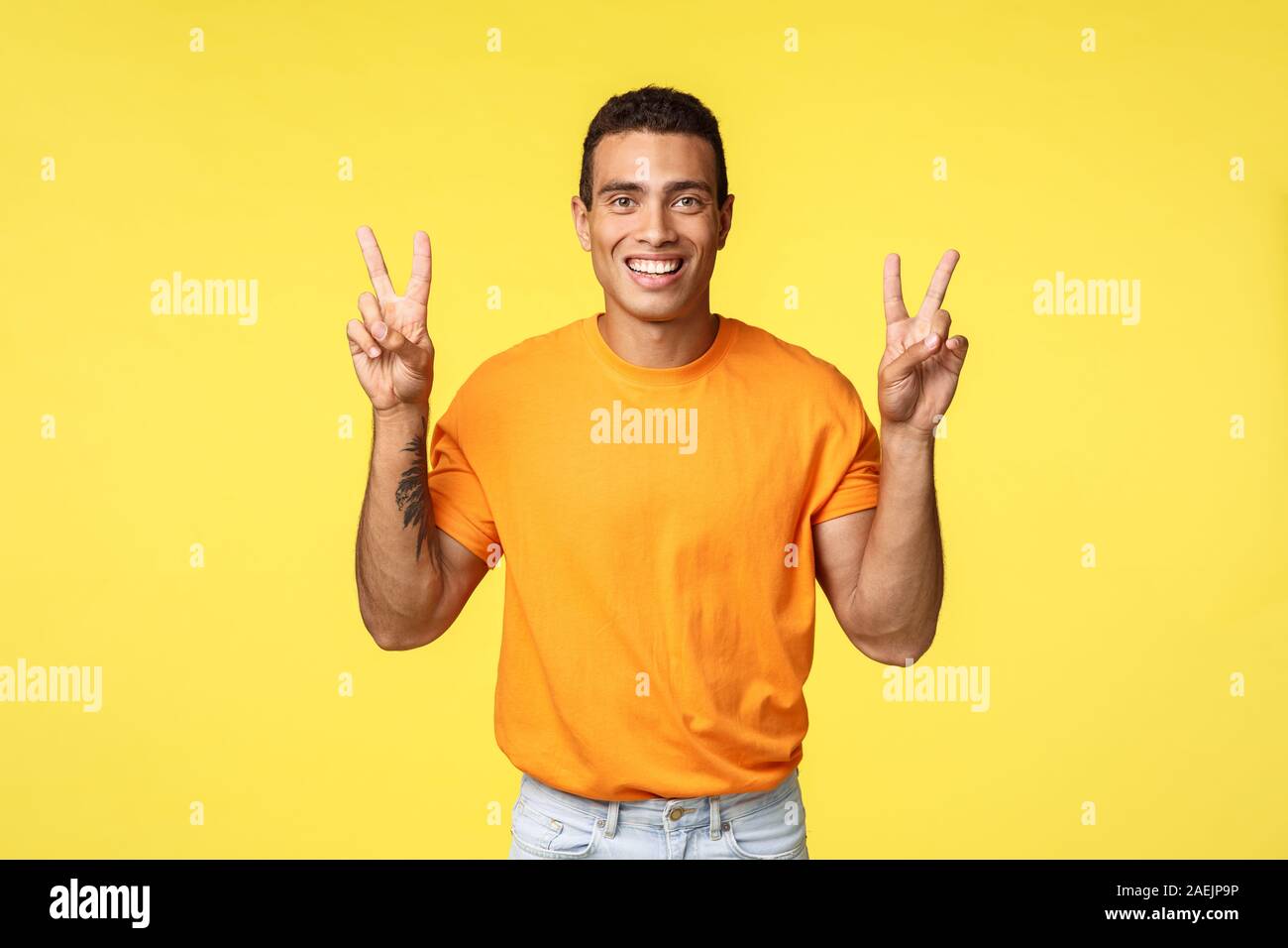 Beau jeune homme joyeux en tee-shirt orange, souriant optimiste, faire la paix geste ou signe citations, fond jaune permanent, la positivité et l'express Banque D'Images