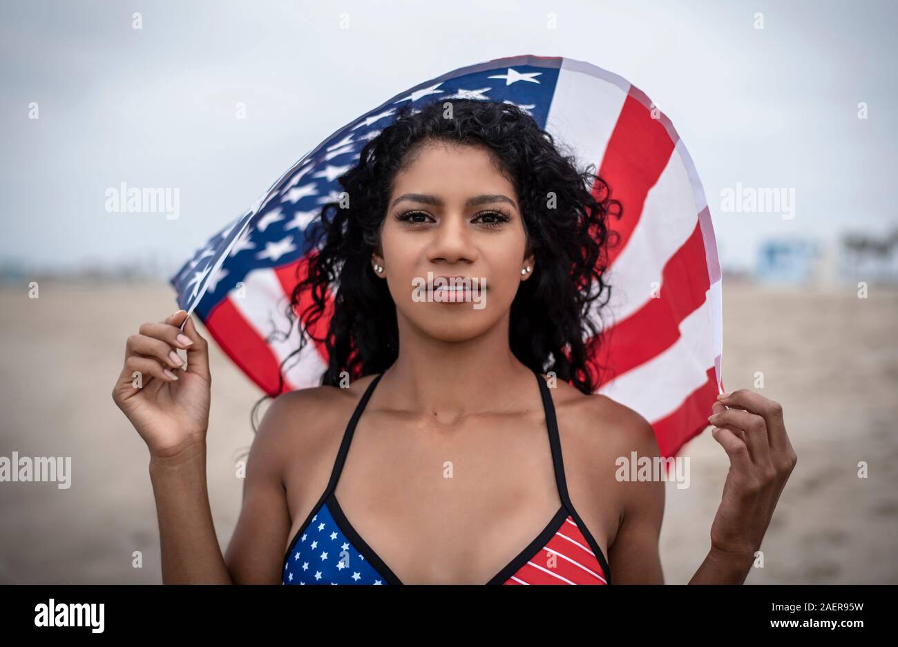 Dark haired Latina détient drapeau américain au-dessus de sa tête tout en portant des Stars and Stripes bikini à la plage le 4 juillet Banque D'Images