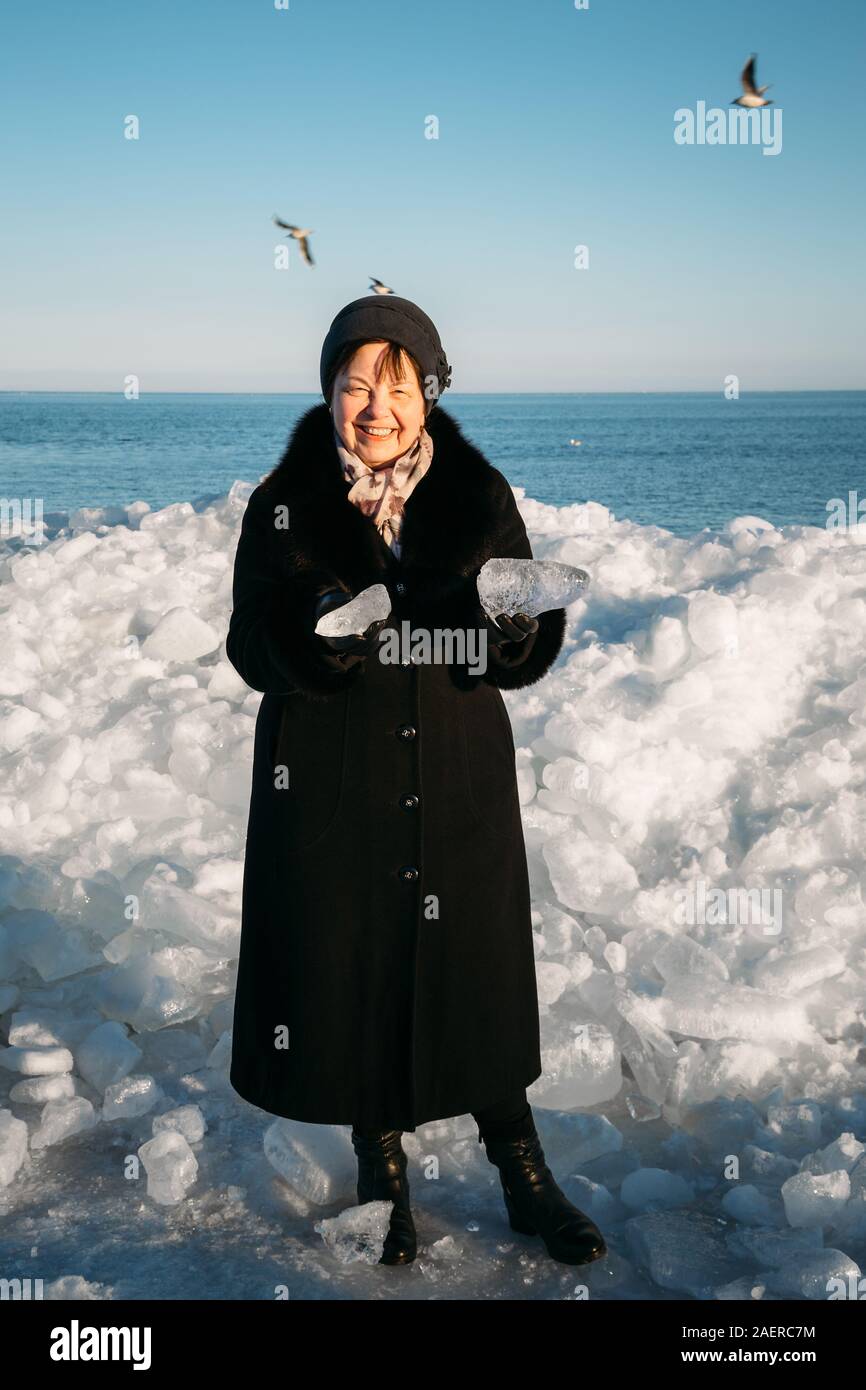Smiling Senior woman in black coat holding morceaux cassés de la glace de mer debout devant la glace en bosses Banque D'Images