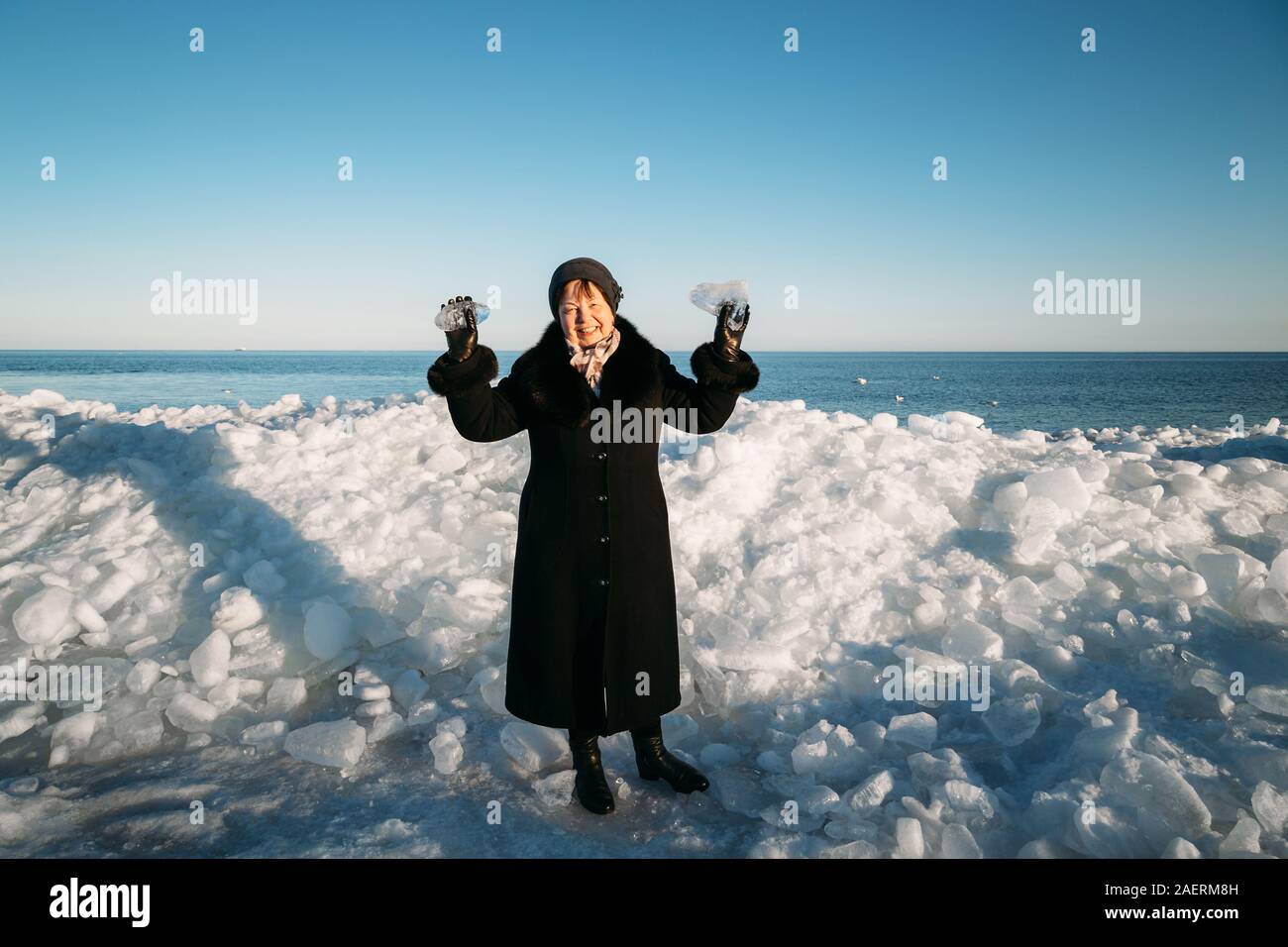 Smiling Senior woman in black coat holding morceaux cassés de la glace de mer debout devant la glace en bosses Banque D'Images