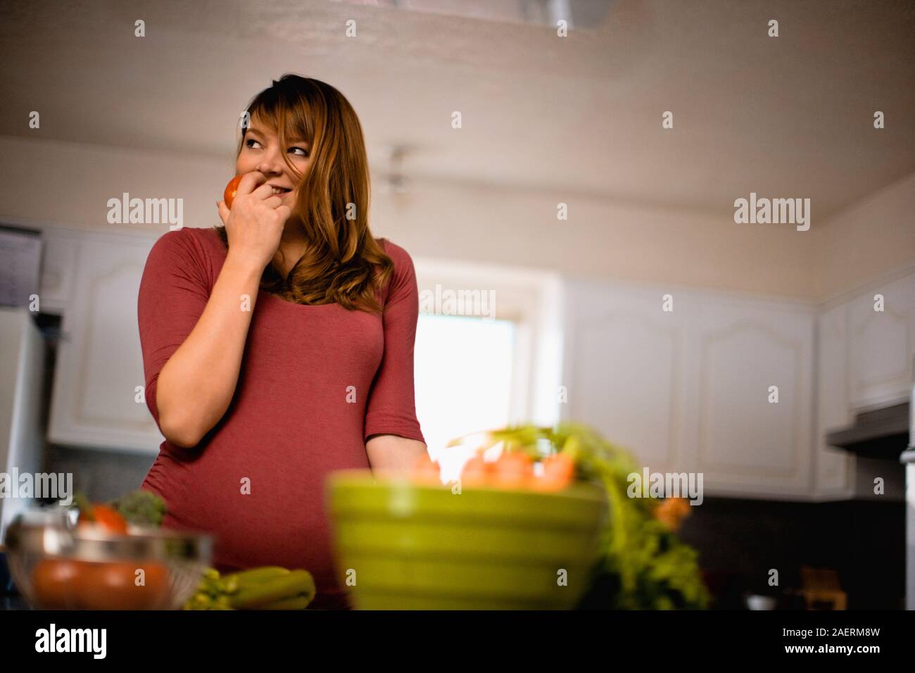 Smiling pregnant woman dans sa cuisine sur le point de prendre une bouchée d'une tomate. Banque D'Images