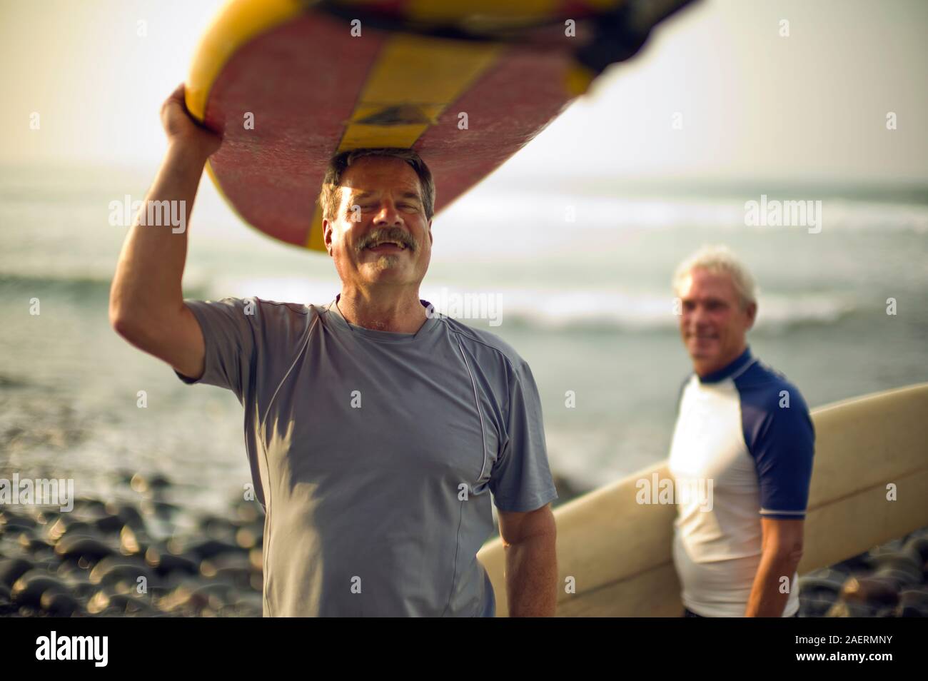 Young man carrying surfboard un rire et son ami masculin transportant un surf sous le bras sourire alors qu'ils posent pour un portrait sur une plage. Banque D'Images