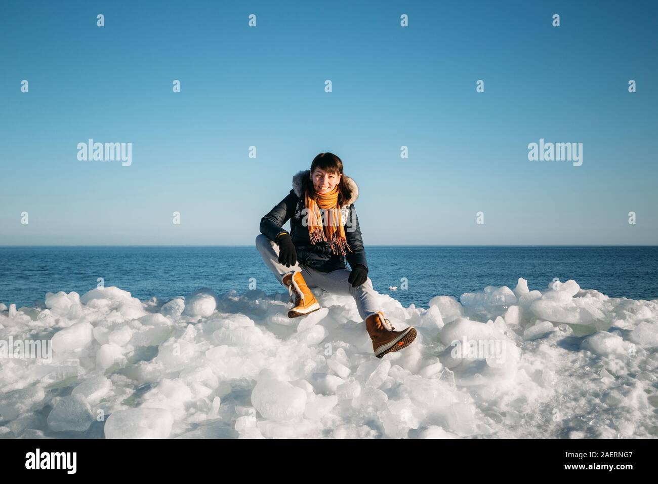 Young smiling woman sitting on a haut de la glace de mer congelés blocs sur une côte de la mer, avec un ciel bleu à l'arrière-plan, à frosty journée ensoleillée Banque D'Images